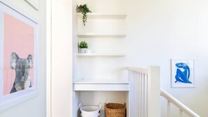 A narrow hallway with white walls, a white door, and a white railing. There are two baskets on the floor, a framed picture on the left, and a framed artwork on the right. - Oversized Artdeco Apartment w 4m high ceilings (Glebe)