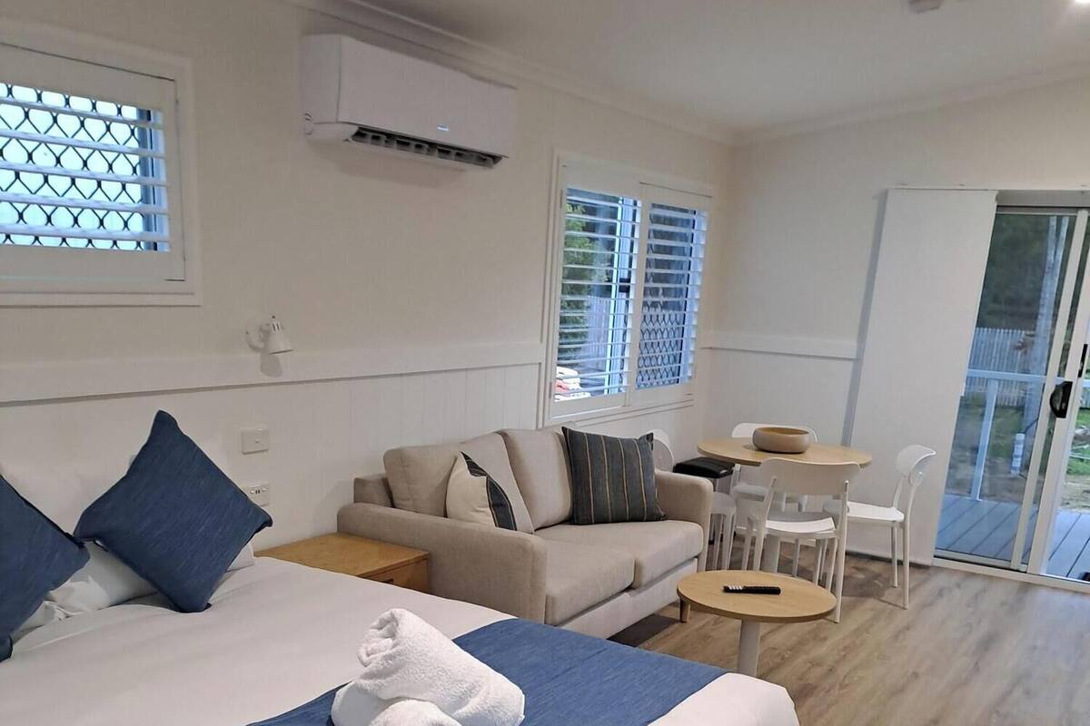 Habitación de hotel con cama, sofá, una mesita y una silla. Tiene una ventana con persianas y un equipo de aire acondicionado en la pared.