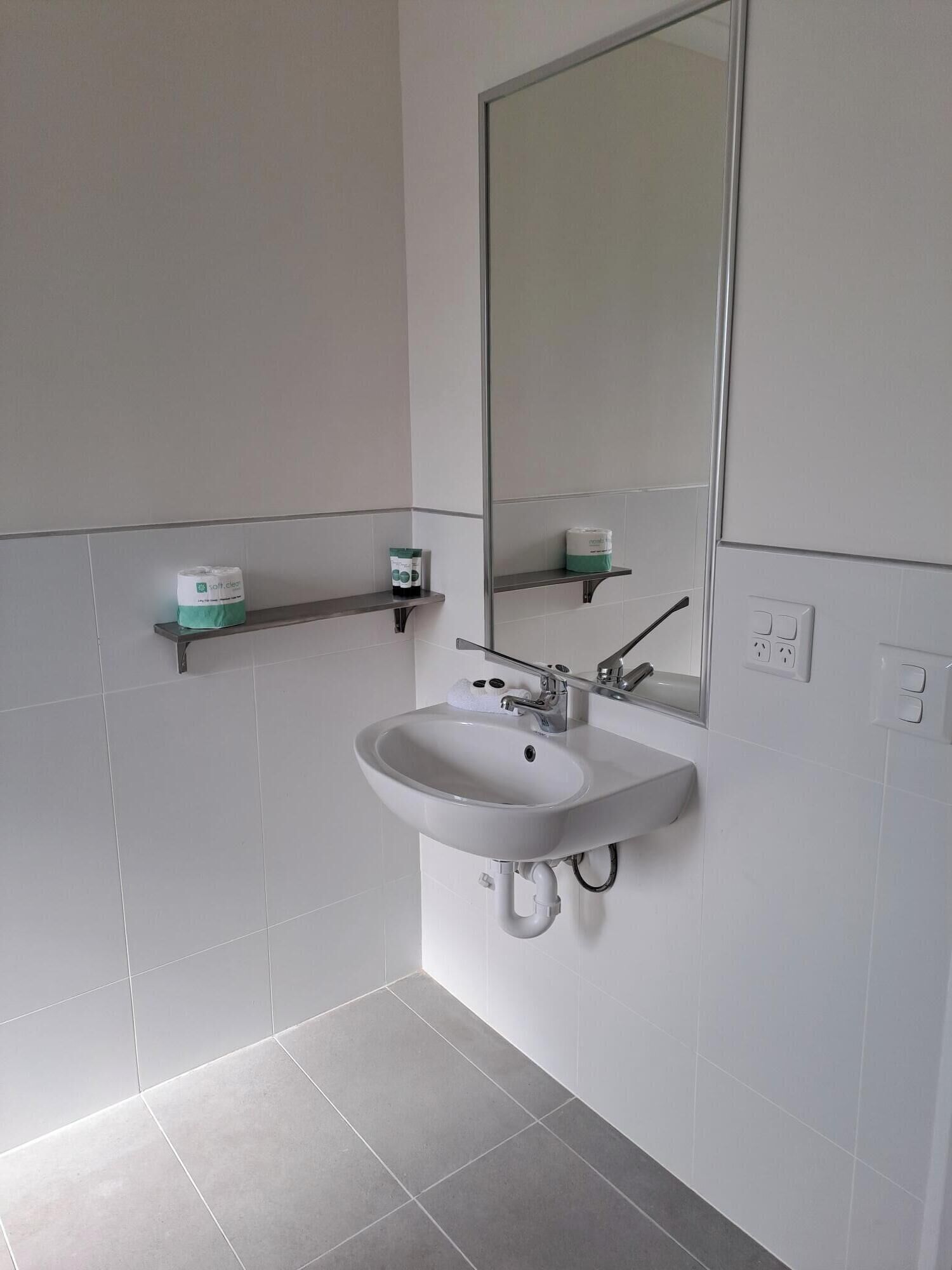 Un baño con lavamanos blanco, espejo y dos repisas con artículos de tocador.