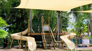 Eine Rutsche mit zwei Gleitbahnen, eine Holzplattform und ein Poolbereich, umgeben von Bäumen.