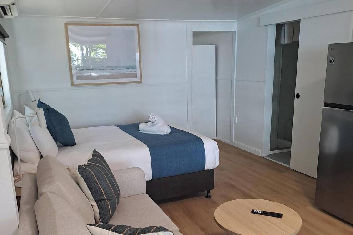 Una habitación moderna y bien iluminada con una cama, un sofá, una pequeña mesa redonda y un refrigerador.