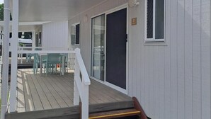 Eine überdachte Terrasse mit einer Holztreppe, die zu einer Glastür führt, einem weißen Geländer und Blick auf einen Patio-Bereich mit grünen Stühlen.