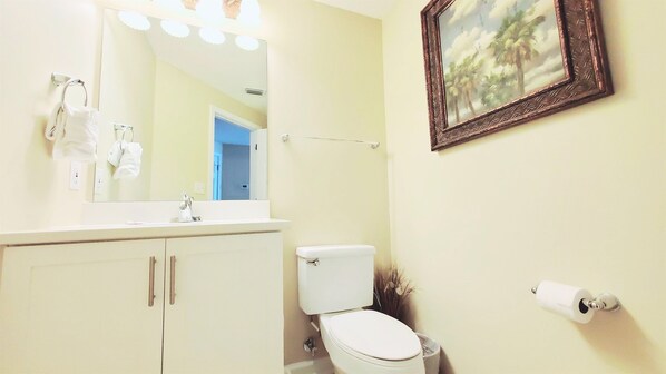 A bathroom with a toilet, sink, and mirror. There is a framed picture on the wall and a roll of toilet paper on the floor. - Bristol 1002 Beach Club-Direct Gulf View-Right On The Beach-Pool/Hot Tub-WiFi! (Gulf Shores)