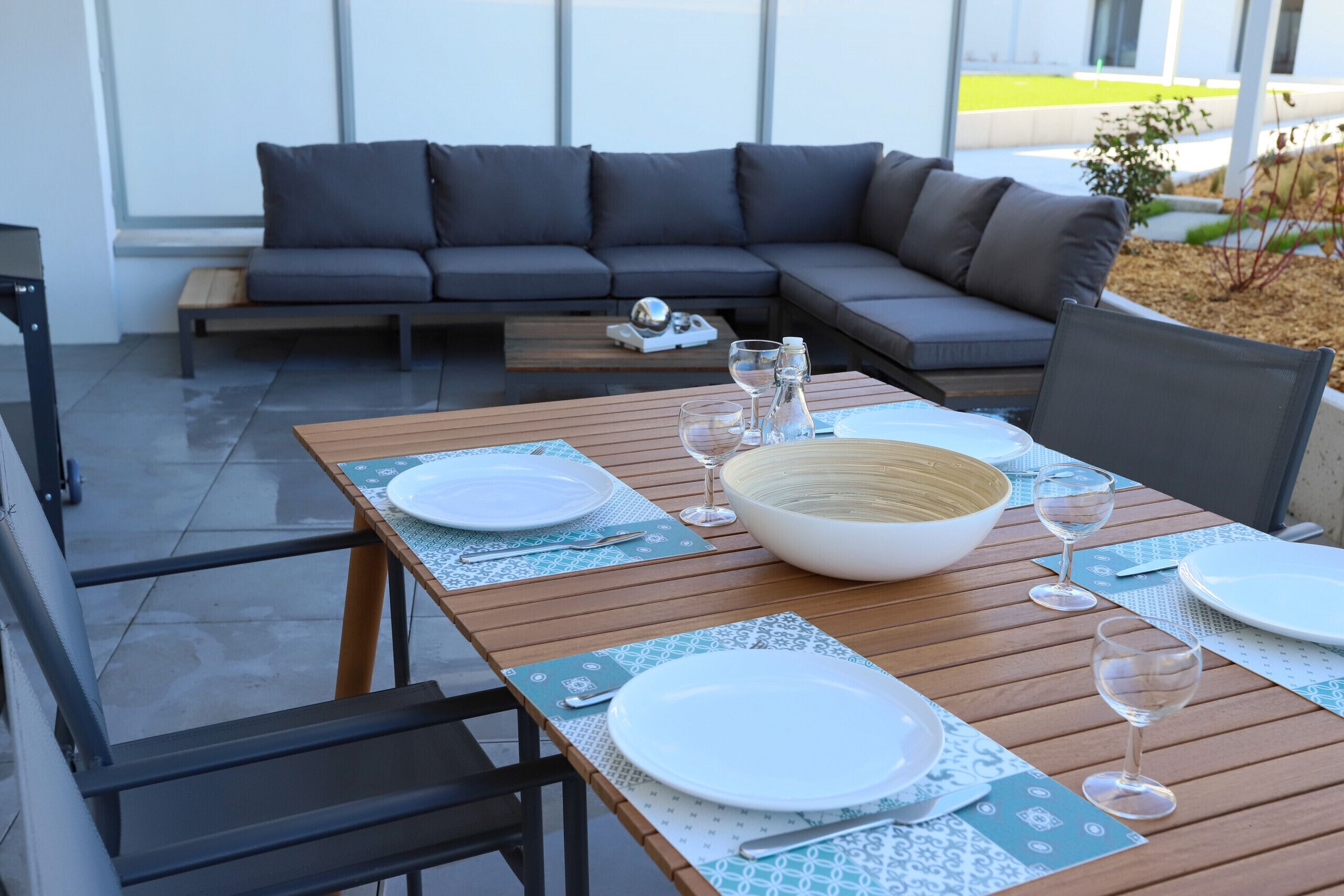 A dining area with a wooden table set for two, a gray sofa, and a view of a green landscape through large windows.