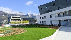 A modern building with a flat roof and large windows, surrounded by a well-maintained lawn and a mountain backdrop. - Reflet du Parmelan Annecy (Annecy)