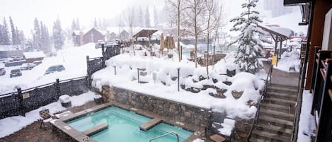 Una piscina con pared de piedra y plataforma de madera, rodeada de suelo y árboles cubiertos de nieve.