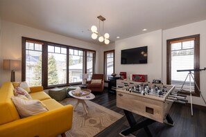 Ein modernes Wohnzimmer mit einem gelben Sofa, einem Kicker, einem Fernseher und großen Fenstern.