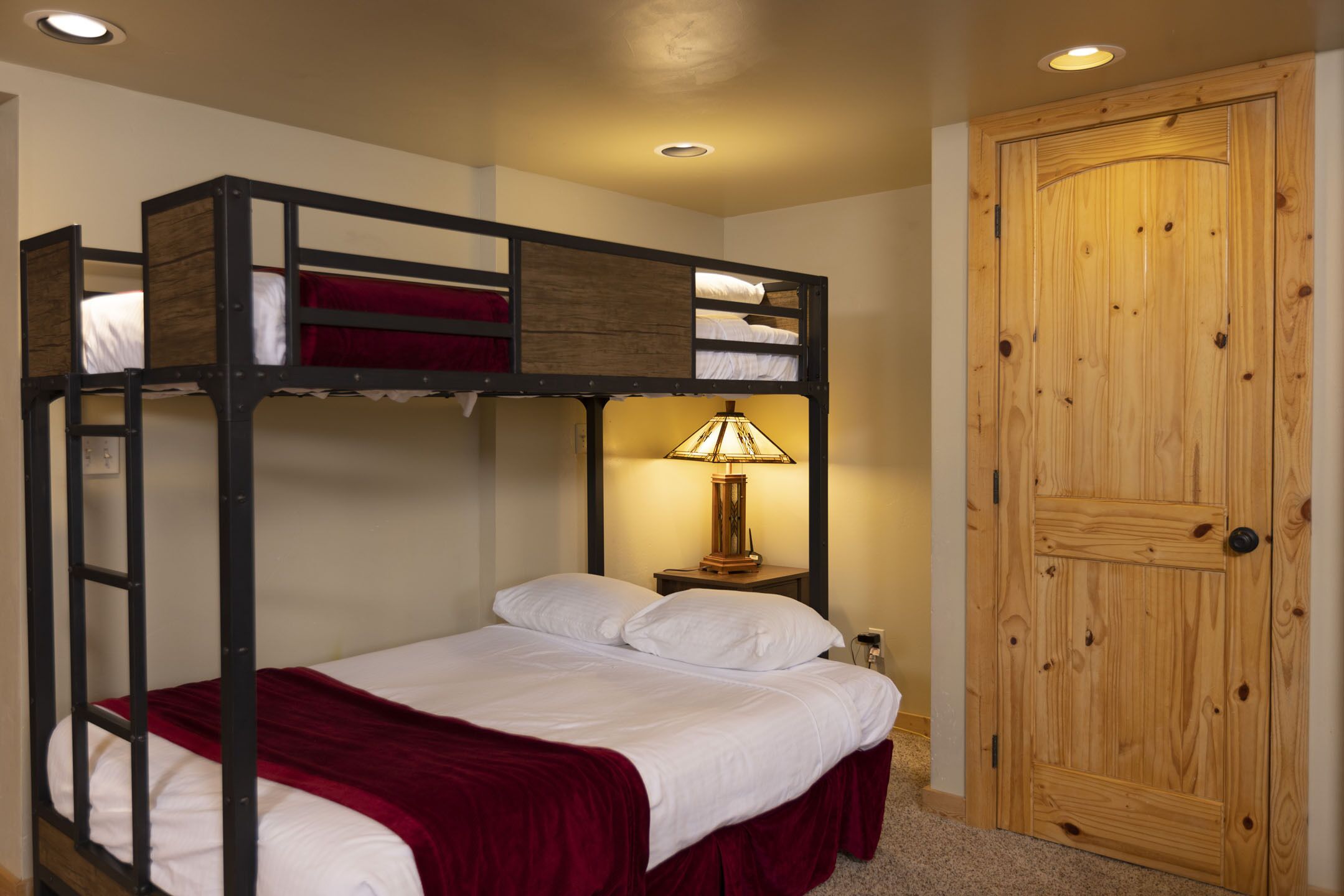 A bunk bed with a red blanket, a wooden nightstand with a lamp, and a wooden door.
