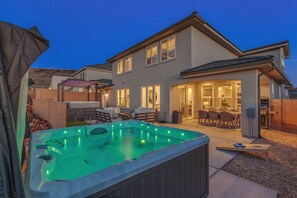 A two-story house with a hot tub in the backyard, a covered patio, and a well-lit interior.