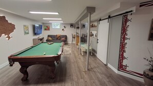 Taula de bitlles amb superfície de feltre verd, un sofà amb coixins, un televisor fixat a la paret i un llit amb capçalera blanca.