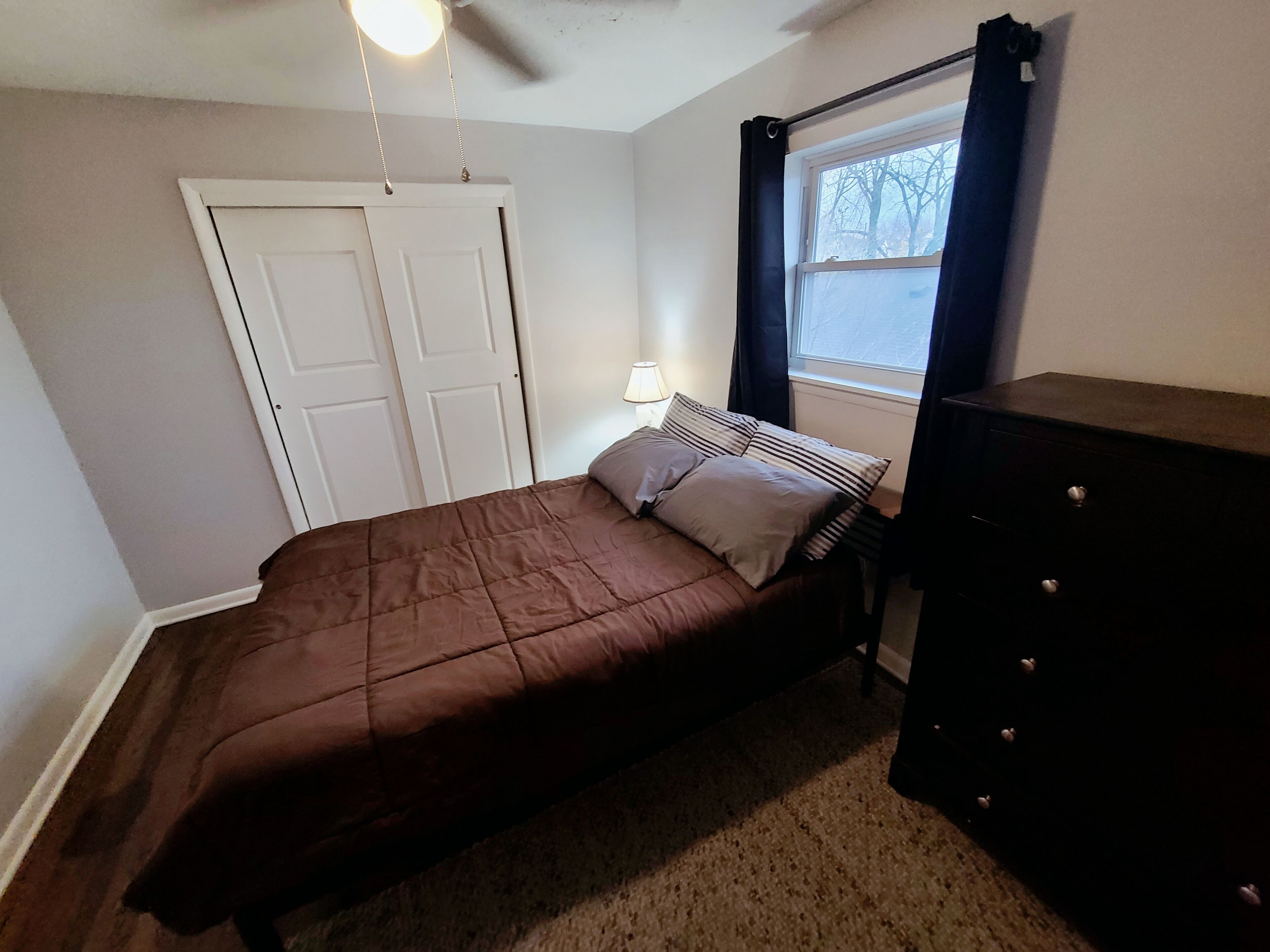 A bedroom with a bed, a dresser, a window with curtains, a ceiling fan, and a door.