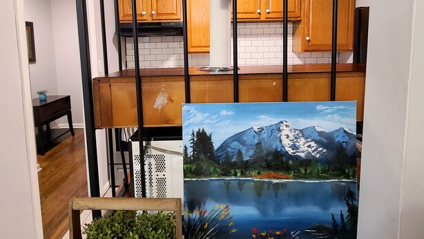 A kitchen with wooden cabinets, a black metal rack, a painting of a mountain landscape, and a sign that reads "MAGIC IS SOMETHING YOU MAKE". - Central Stay! Near UK/Downtown/Campus. (Lexington)