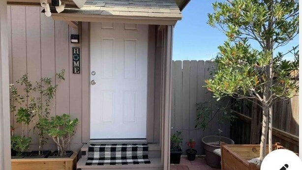 Une porte d’entrée avec une plaque portant l’inscription « MAISON », un paillasson à carreaux et des plantes en pot.