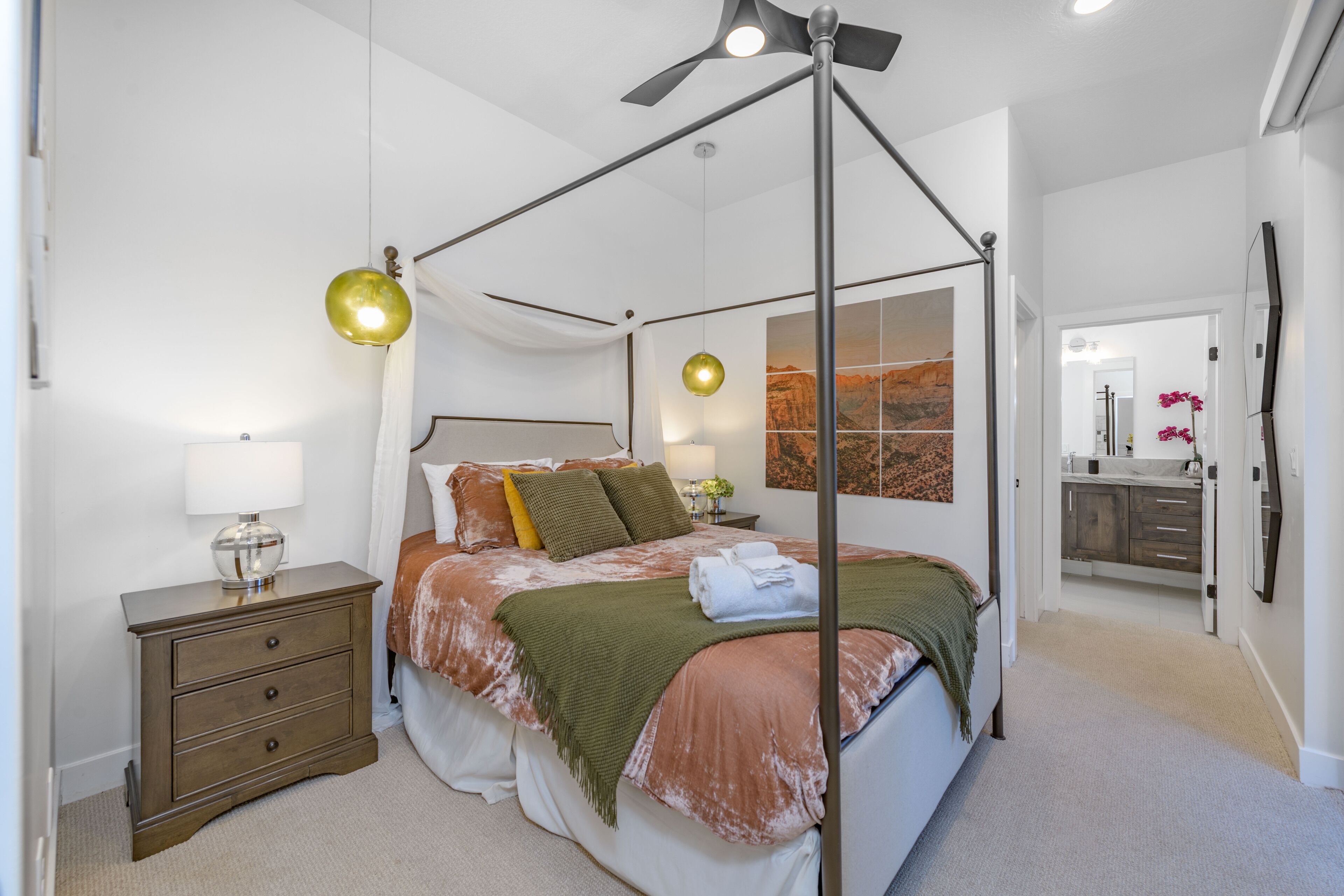 A modern bedroom with a four-poster bed, a bedside table with a lamp, and a view into the bathroom.