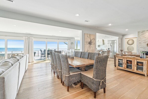 A spacious dining area with a large wooden table and wicker chairs, a built-in wooden cabinet, and a view of the ocean through large windows.
