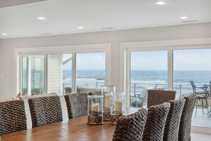 A dining area with a wooden table, wicker chairs, and a view of the ocean through large windows. - White Sands | Luxury Oceanfront Home | Direct Beach Access | Expansive Deck + Chef's Kitchen (Folly Beach)