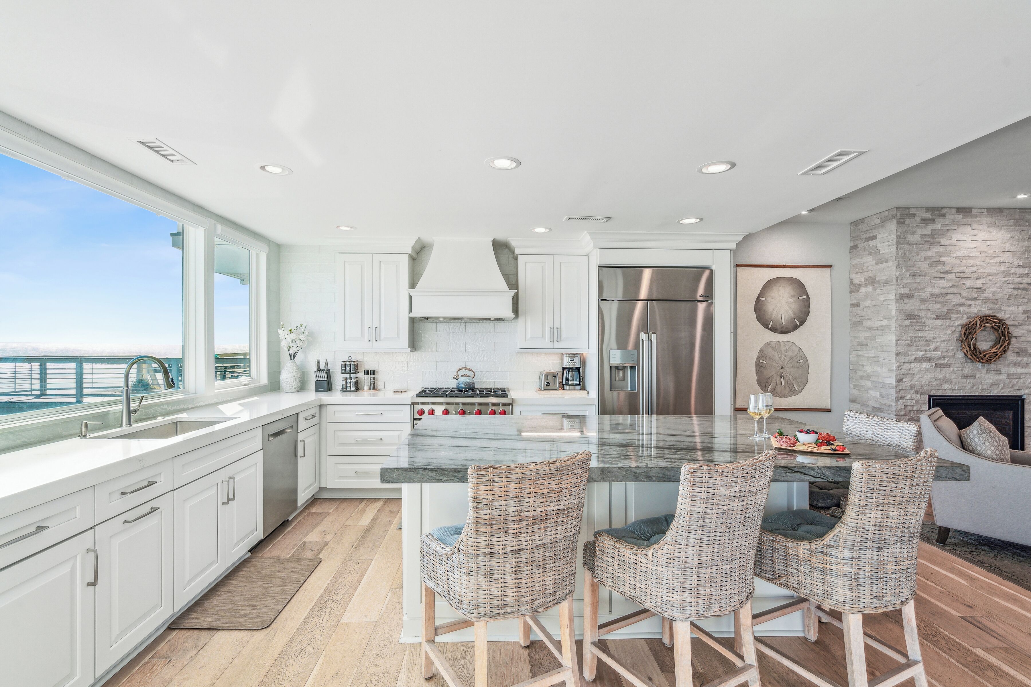 A modern kitchen with white cabinets, a large island with bar stools, a stainless steel refrigerator, and a view of the ocean.