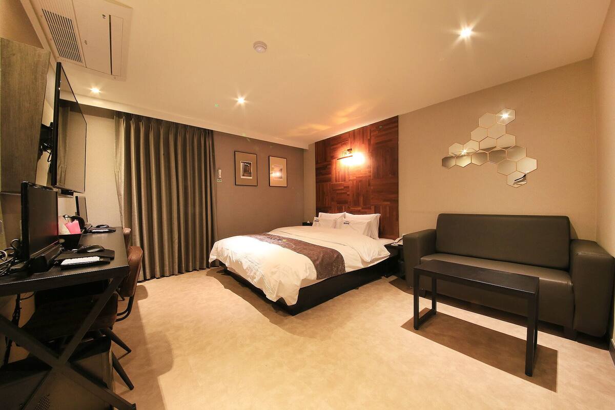 A bedroom with a bed, a desk, a chair, a sofa, and a wall-mounted light fixture.