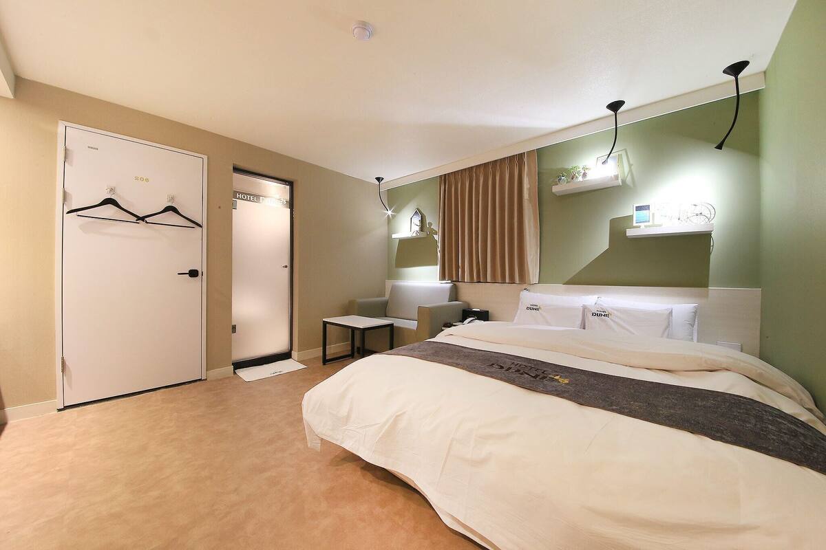 A hotel room with a bed, a desk, a chair, a coat rack, and a door.