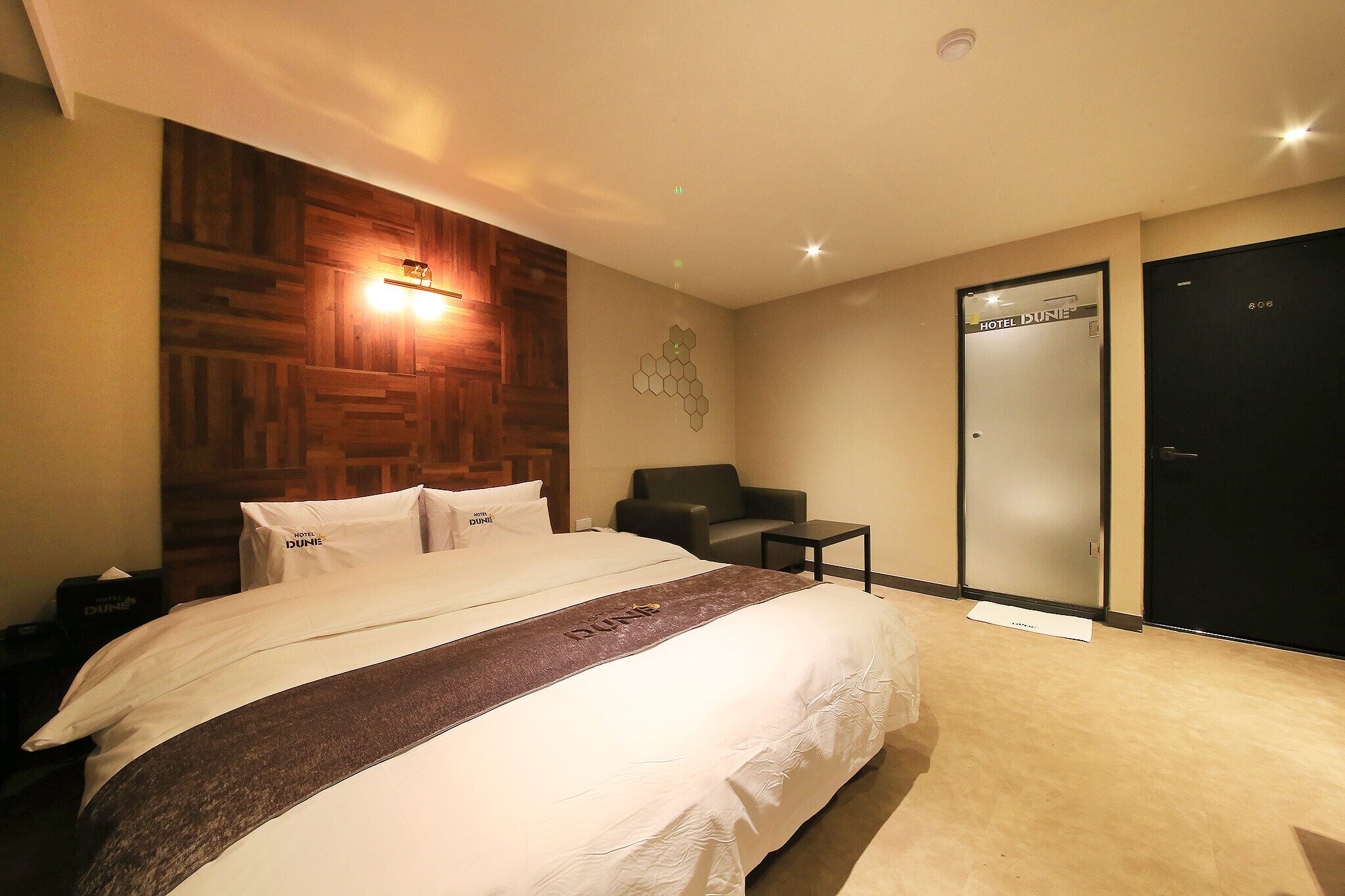 A hotel room with a large bed, a wooden headboard, a wall-mounted light, a chair, a small table, and a door labeled "HOTEL DUNE".