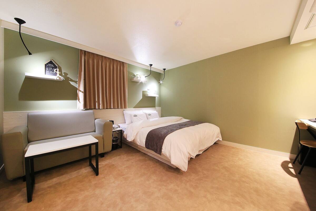 A bedroom with a bed, a desk, a chair, a bench, and a hanging light fixture.