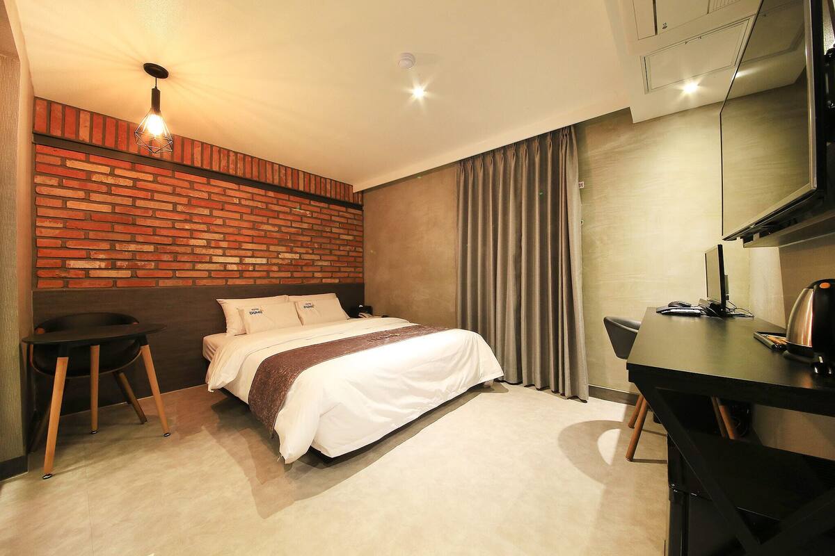 A hotel room with a brick accent wall, a bed with white bedding, a desk with a kettle, and a chair.
