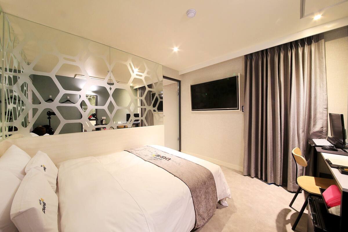 A hotel room with a bed, a desk with a chair, a TV, and a curtain.