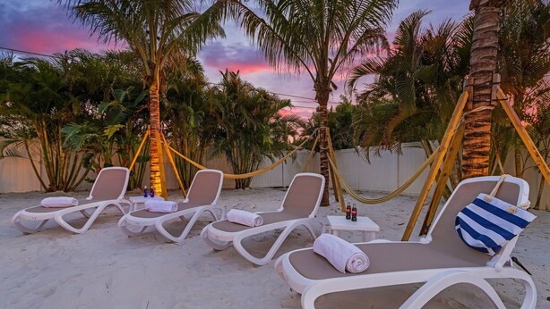 Un área de descanso frente a la playa con sillas reclinables blancas, una hamaca y vistas a palmeras bajo un cielo al atardecer.