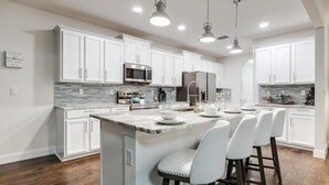 Una cocina moderna con armarios blancos, una isla grande con taburetes de bar, electrodomésticos de acero inoxidable e iluminación colgante.