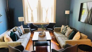 A living room with two sofas, a coffee table, and two armchairs. There is a large window with natural light and a flat-screen TV mounted on the wall.