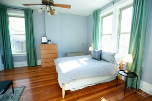 A bedroom with a wooden floor, a bed with a white and blue striped bedspread, a dresser, a nightstand with a lamp, and a ceiling fan. - The Convent (Cincinnati)