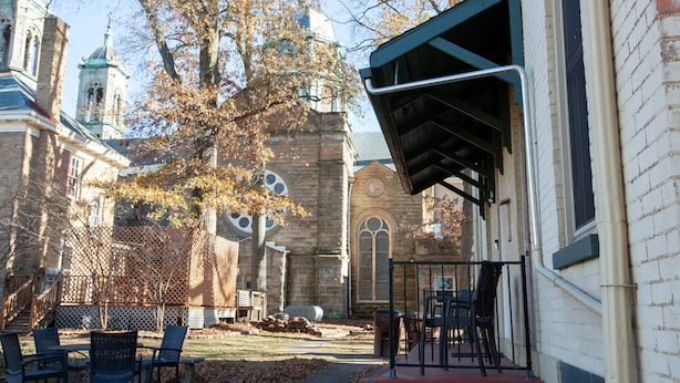 An outdoor seating area with tables and chairs, a brick building with a green awning, and a large tree with autumn leaves.