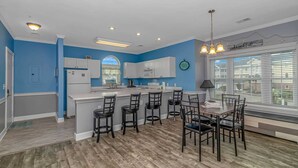 A modern kitchen with a central island, bar stools, a dining table, and a large window with blinds. - Magnolia N 201 (Myrtle Beach)