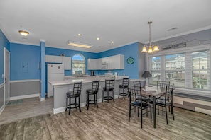 A modern kitchen with a central island, bar stools, a dining table, and a large window with blinds.