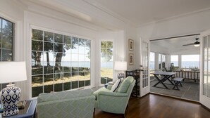 Amplia sala de estar con grandes ventanales que dan a una playa, zona de comedor con mesa y sillas, y un rincón de descanso acogedor con un sillón estampado.