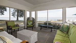 Habitación luminosa con grandes ventanales que dan a una playa, amueblada con mobiliario de mimbre y un banco.