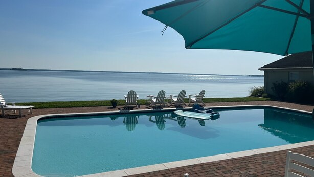 Piscina con vista al agua, rodeada de sillones de playa y un gran parasol.