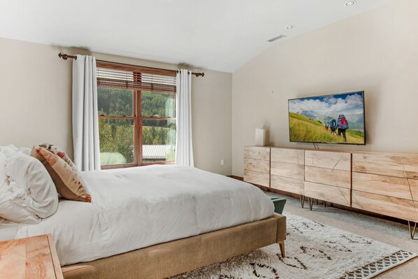 Un dormitorio con una cama grande, un aparador de madera, un televisor en la pared y una ventana con persianas.