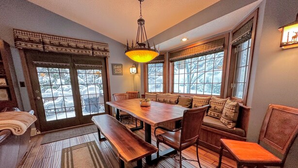 A cozy dining area with a wooden table and benches, large windows with snow outside, and a warm hanging light fixture. - FC17 Stylishly renovated Forest Cottage, walk to ski trails, wifi, cable, ping pong, AC! (Bretton Woods)