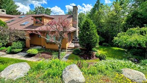 A house with a stone chimney, a covered patio, and a well-maintained garden with various plants and a lawn. - FC17 Stylishly renovated Forest Cottage, walk to ski trails, wifi, cable, ping pong, AC! (Bretton Woods)