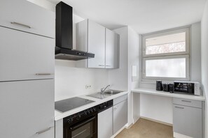 A compact kitchen with white cabinets, a black range hood, a built-in oven, a stovetop, and a window with blinds. - Modern Leopoldstadt 1BR w/ Balcony & Elevator, nr metro U2, by Blueground (Vienna)