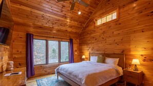 Un dormitorio de cabaña de madera con cama, mesitas de noche, una ventana con cortinas y un ventilador de techo.