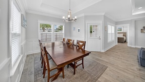 Un vaste espace dédié à la salle à manger, avec une grande table et des chaises en bois, un lustre et de grandes fenêtres équipées de stores.