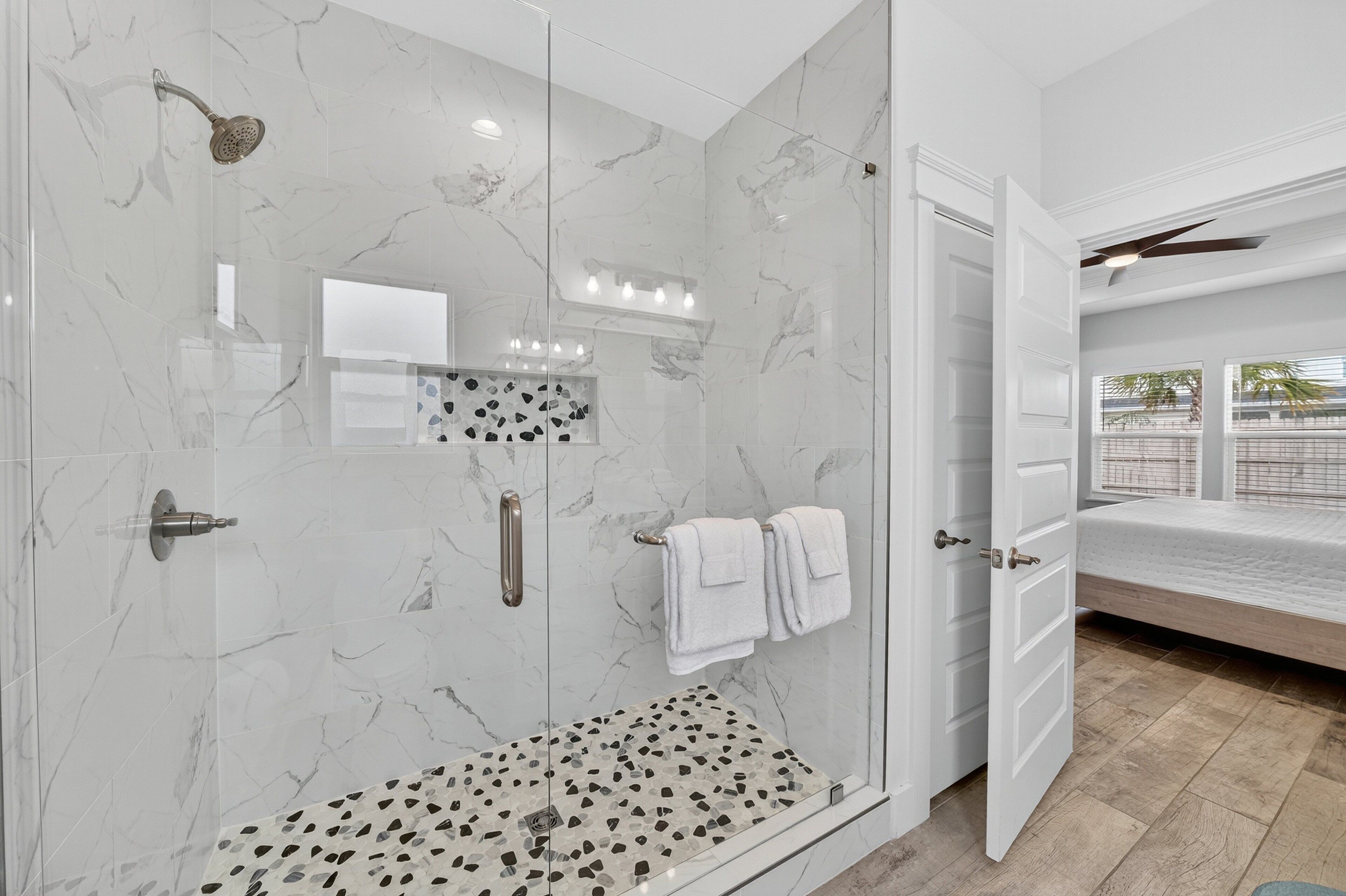 A modern bathroom with a glass-enclosed shower, marble walls, and a visible bed in the adjacent room.