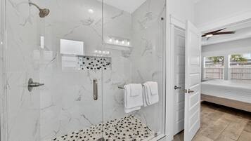 A modern bathroom with a glass-enclosed shower, marble walls, and a visible bed in the adjacent room.
