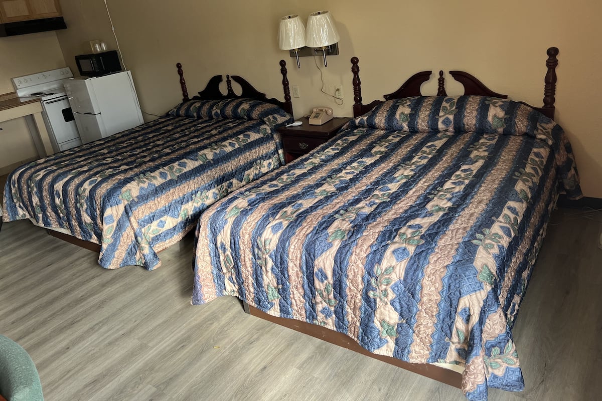 Two beds with patterned bedding, a small kitchenette with a microwave and stove, and a nightstand with a lamp.