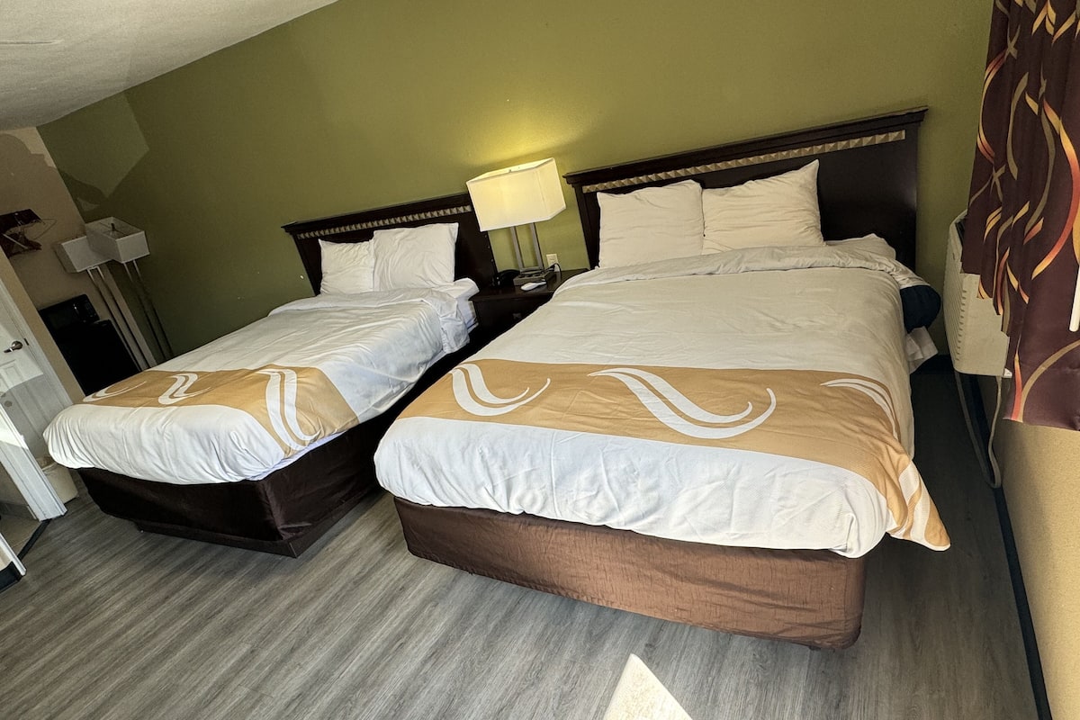 A hotel room with two beds, a nightstand with a lamp, and a door.