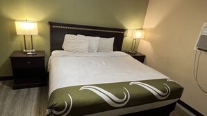 A hotel room with a bed, two bedside tables with lamps, a wall-mounted air conditioner, and a wooden floor. - Country Inn-Giddings (Giddings)