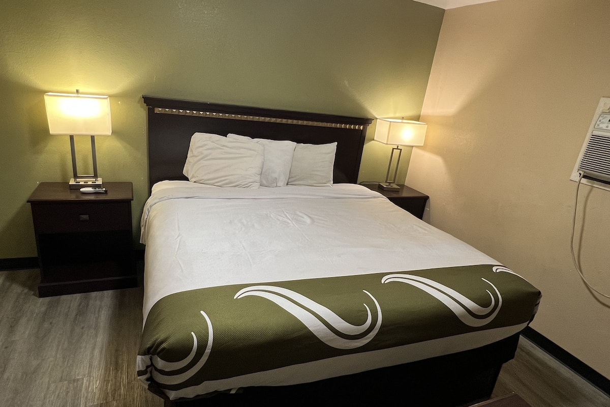 A hotel room with a bed, two bedside tables with lamps, a wall-mounted air conditioner, and a wooden floor.