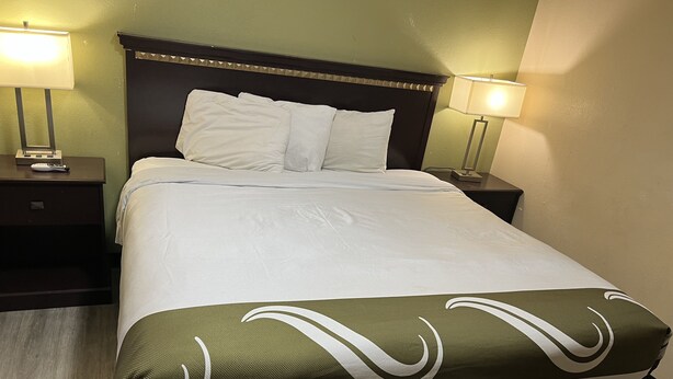 A neatly made bed with white linens and a green and white patterned throw. There are two bedside tables with lamps, and the room has a wooden floor. - Country Inn-Giddings (Giddings)
